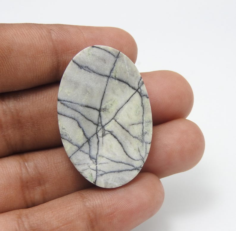 May include: Oval-shaped gemstone with a light gray and green base, crisscrossed by dark gray veins. The stone is held in a hand, showcasing its polished surface and natural pattern. A unique piece for jewelry or collection.