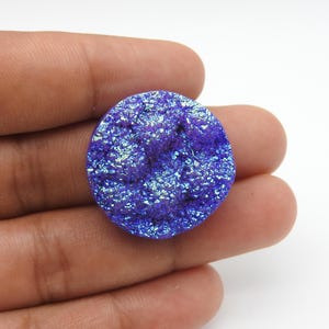 Puede incluir: Una gema redonda y brillante, de color azul-púrpura, con una superficie texturizada. La gema tiene una apariencia cristalina con reflejos iridiscentes. La piedra se sostiene en una mano sobre un fondo blanco. Esta gema podría usarse para la fabricación de joyas.