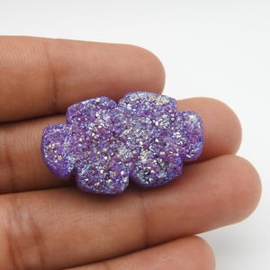 Puede incluir: Primer plano de un cabujón de piedra preciosa druzy púrpura con forma de flor. La piedra preciosa tiene una superficie cristalina brillante con tonos de púrpura y plata. El cabujón se sostiene en una mano sobre un fondo blanco.