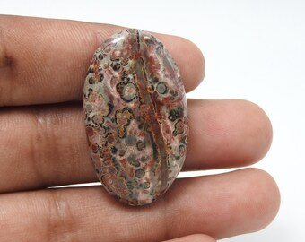 May include: An oval-shaped gemstone with a speckled pattern in shades of red, brown, green, and black. The polished surface of the stone displays a natural, organic design. The gemstone is held in a hand.