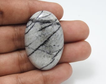 Puede incluir: Gema ovalada con apariencia veteada. La piedra presenta una base de tonos blancos y grises, atravesada por audaces vetas negras. La superficie pulida refleja la luz, resaltando los patrones naturales. La gema se sostiene en una mano, mostrando su tamaño y forma.
