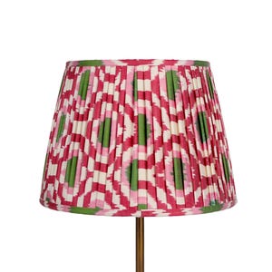 May include: A lampshade with a pleated design featuring a pattern of red, green, and white shapes. The lampshade is mounted on a brass-coloured metal stand. The overall design is decorative and colourful.