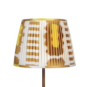 May include: A pleated lampshade with a white background and a pattern of yellow and brown shapes. The lampshade is cone-shaped and sits atop a thin, gold-colored metal stand. The lamp is against a white background.