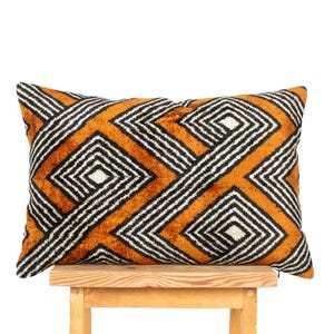 May include: A rectangular throw pillow featuring a vibrant burnt orange velvet texture with a repeating geometric pattern of black and white concentric diamonds and intersecting lines that span across the entire surface of the fabric cover.