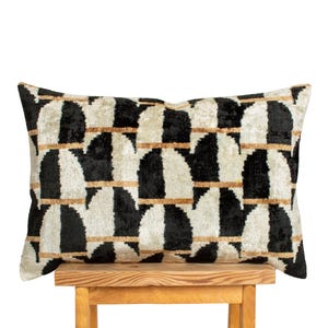 May include: Rectangular decorative pillow with a geometric pattern. The pillow features a repeating design of black and cream-colored semi-circles, connected by thin, horizontal gold lines. The pillow is resting on a small wooden stool.