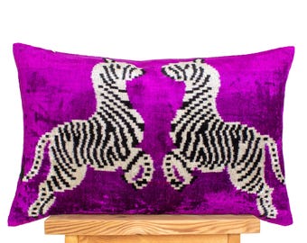 Op de afbeelding: Een rechthoekig sierkussen met een levendige magenta achtergrond. Twee zebra's met zwart-witte strepen staan op het kussen. Het kussen ligt op een lichtbruine houten kruk.