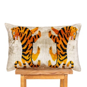 May include: Rectangular cushion featuring two stylized tigers in orange and black stripes against a light beige background. The tigers are in a playful pose, with their arms raised. The cushion is resting on a wooden stool.