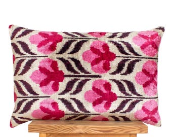 Könnte beinhalten: Ein rechteckiges Zierkissen mit floralem Muster. Das Kissen hat einen cremefarbenen Hintergrund mit einem sich wiederholenden Muster aus rosa und dunkelroten Blumen und Blättern. Es liegt auf einem Holzschemel.
