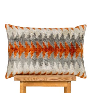 May include: Rectangular cushion with a geometric pattern. The cushion has a cream background with a repeating zigzag design in grey and orange. It is resting on a wooden stool.