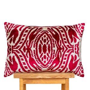 Pode incluir: Uma almofada decorativa retangular com um padrão vermelho e creme. A almofada está colocada sobre um banco de madeira. O padrão é um design simétrico com uma textura aveludada. Mede cerca de 60 cm de comprimento.