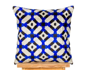 May include: Square cushion with a geometric pattern in black, white, and blue. The cushion is placed on a small wooden stool. The pattern features diamond shapes and intersecting lines, creating a modern design. The cushion is approximately 40cm x 40cm.