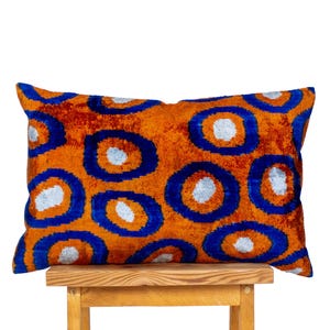 May include: An orange rectangular pillow with a repeating pattern of blue circles with white centers. The pillow is made of a plush, textured fabric and rests on a wooden stool.