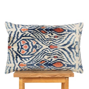 Op de afbeelding: Een rechthoekig sierkussen met een symmetrisch ikat-patroon in tinten diep marineblauw, gebrand oranje en crème. De stof heeft een zachte, fluweelachtige textuur met een herhalend abstract ontwerp dat het gehele oppervlak van het kussen beslaat.