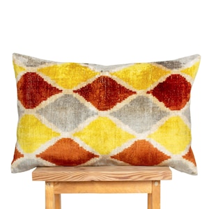 May include: A rectangular decorative pillow with a geometric diamond pattern in shades of yellow, orange, and gray. The pillow is placed on a small wooden stool against a white background. The pillow's design adds a touch of color and texture.