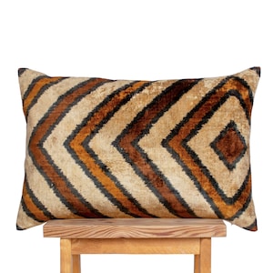 May include: Rectangular decorative pillow with a geometric pattern. The pillow features a repeating chevron design in shades of brown, orange, and black on a beige background. The pillow is resting on a wooden stool.