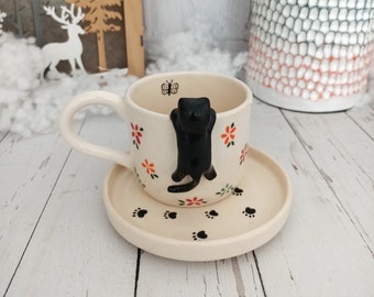 May include: A cream-colored ceramic teacup and saucer set. The cup features a black cat climbing the side, with floral and paw print details. A small butterfly is also on the cup. The saucer has paw prints.