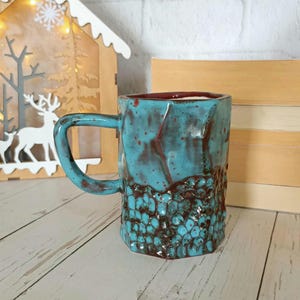 Puede incluir: Taza de cerámica turquesa hecha a mano, con una base texturizada marrón. La taza tiene forma rectangular con asa e interior rojo oscuro. La taza se exhibe sobre una superficie de madera clara.