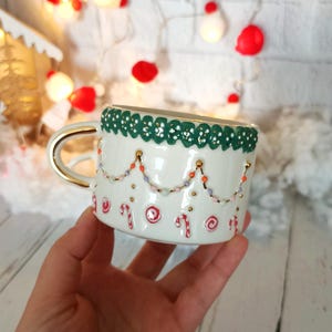 Puede incluir: Taza de cerámica blanca con borde verde y un diseño festivo de bastones de caramelo y una guirnalda de cuentas.