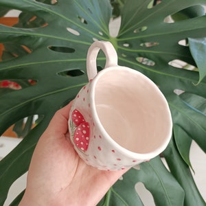 May include: White ceramic mug with a red and white mushroom design. The mug has a handle and is decorated with red polka dots.