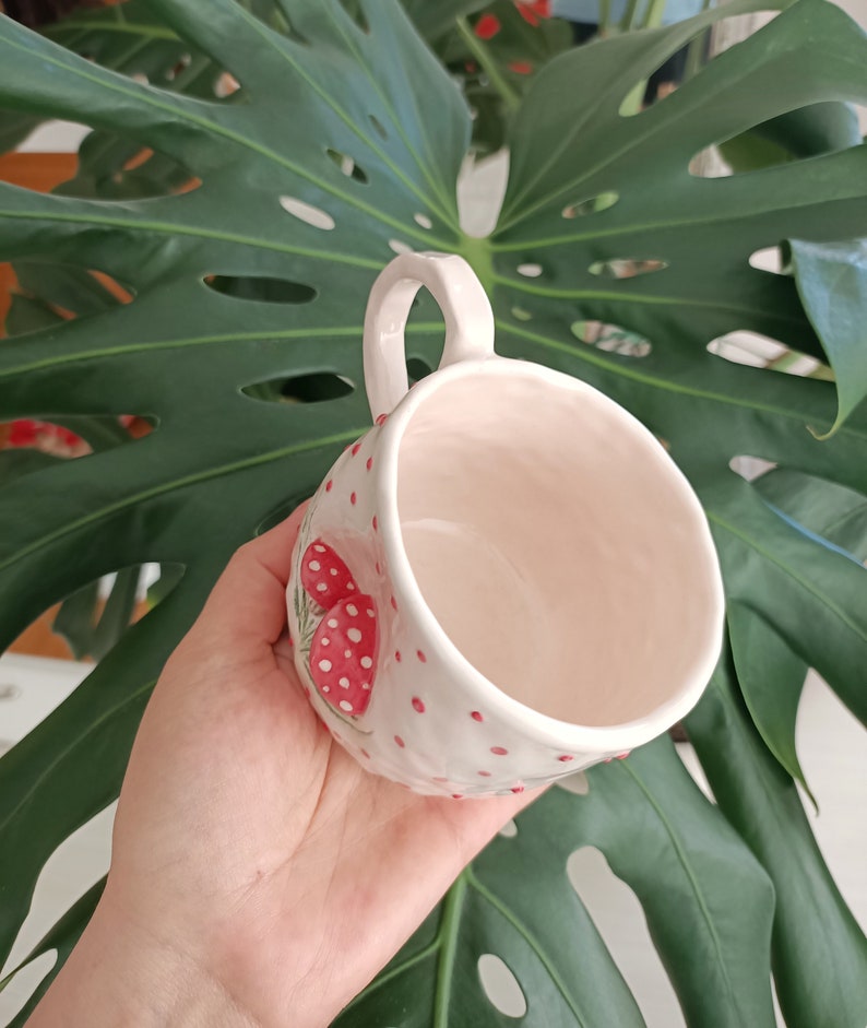 May include: White ceramic mug with a red and white mushroom design. The mug has a handle and is decorated with red polka dots.