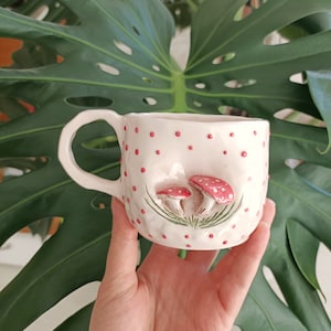 May include: White ceramic mug with a pink and green mushroom design. The mug has a handle and is decorated with red polka dots.