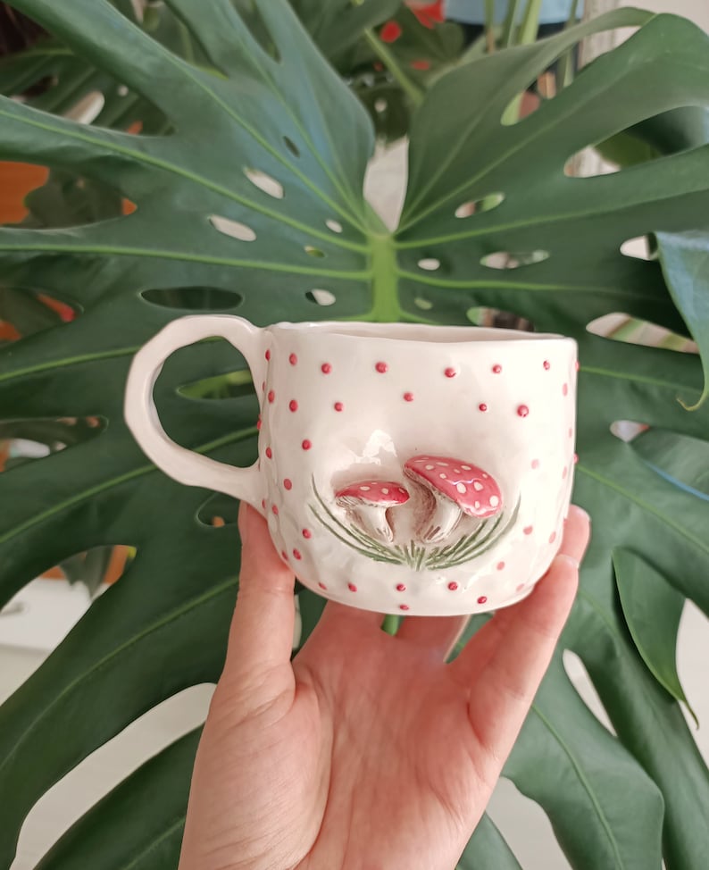 May include: White ceramic mug with a pink and green mushroom design. The mug has a handle and is decorated with red polka dots.