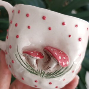 May include: White ceramic mug with red polka dots and a 3D design of two red and white mushrooms with green leaves.