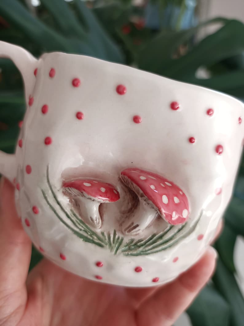May include: White ceramic mug with red polka dots and a 3D design of two red and white mushrooms with green leaves.