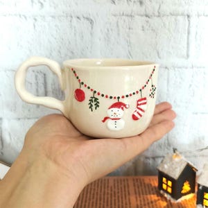 Op de afbeelding: Crèmekleurige keramische mok met een kerstontwerp. De mok heeft een sneeuwpop, rode ornamenten en een kous. De handgreep is groot en gebogen. De mok wordt in een hand gehouden.