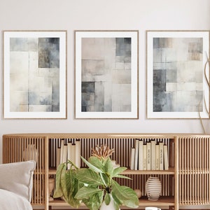 Könnte beinhalten: Drei gerahmte abstrakte Kunstdrucke. Jeder Druck zeigt ein geometrisches Design in Grau-, Beige- und Weißtönen. Das Kunstwerk wird über einem Holzschrank mit Büchern und Dekorationsgegenständen ausgestellt, wodurch eine moderne und minimalistische Ästhetik entsteht.