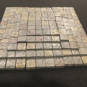 May include: A set of gray and beige stone tiles arranged in a stepped pattern, creating a textured surface. The tiles are designed for use in tabletop games or miniature settings.