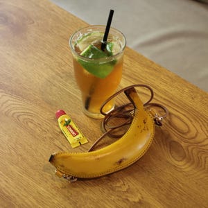 May include: A yellow banana-shaped purse with a brown strap, a glass of iced tea with lime slices and a black straw, and a tube of Carmex lip balm on a wooden surface.
