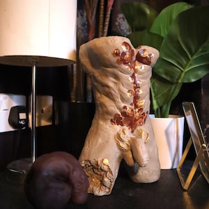 May include: A grey sculpted torso statue with bronze and gold accents. The statue has a detailed depiction of the male anatomy. A dark brown sculpture of a testicle is next to the torso. The statue is on a dark surface.