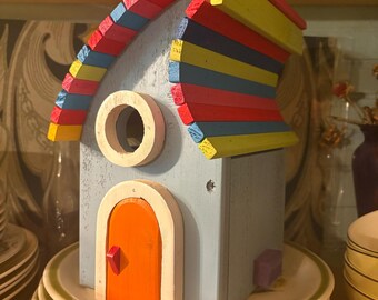 May include: A whimsical, hand-painted wooden birdhouse with a light blue body and a multi-colored roof. The roof features red, yellow, and blue slats. It has an orange door with a red knob and a circular window. The birdhouse is sitting on a stack of plates.