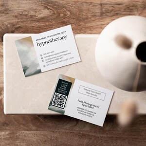 May include: Two white business cards with the text "hypnotherapy" and "Pain Management Specialist". The cards include contact information and a QR code. A white ceramic vase sits in the background.