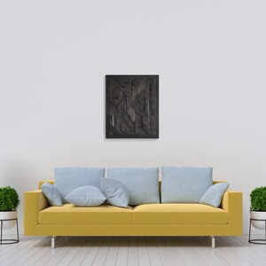 May include: A yellow couch with blue cushions sits in front of a white wall with a black and brown geometric wall art hanging above it. A white lamp with a wooden base and a potted plant are on the left side of the couch. A small wooden table with a potted plant and a white box on top is on the right side of the couch.