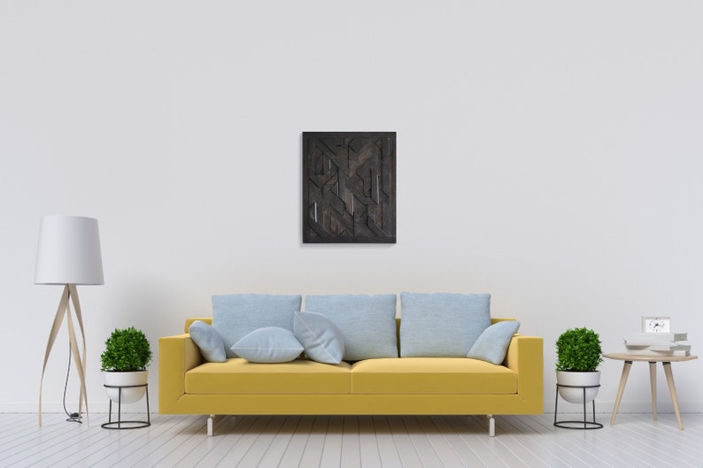 May include: A yellow couch with blue cushions sits in front of a white wall with a black and brown geometric wall art hanging above it. A white lamp with a wooden base and a potted plant are on the left side of the couch. A small wooden table with a potted plant and a white box on top is on the right side of the couch.
