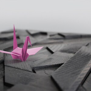 May include: A pink origami crane sits on a dark wood surface with a geometric pattern.