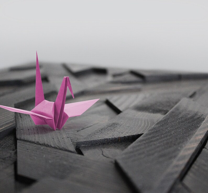 May include: A pink origami crane sits on a dark wood surface with a geometric pattern.