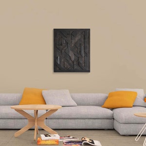 May include: A gray sectional sofa with two orange throw pillows, a wooden coffee table, and a stack of books on a metal side table. A black and white geometric wooden wall art hangs above the sofa.