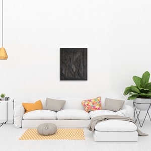 May include: A white sectional sofa with a gray throw blanket and colorful pillows sits in a living room with a black geometric wall art piece, a small round table with a potted plant, and a plant stand with a large green plant.