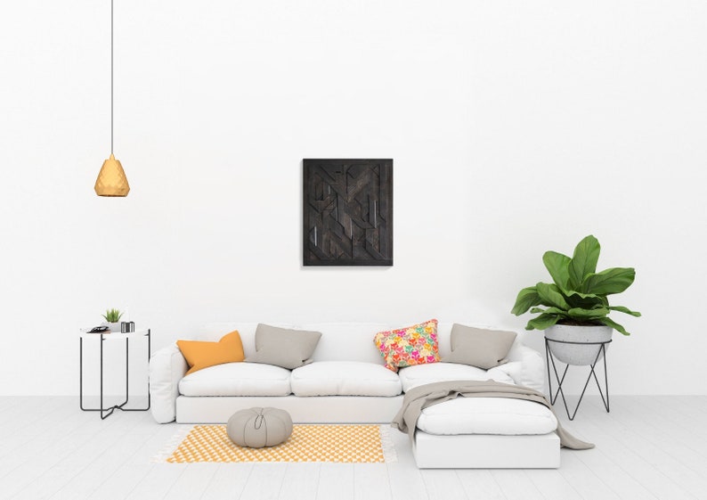 May include: A white sectional sofa with a gray throw blanket and colorful pillows sits in a living room with a black geometric wall art piece, a small round table with a potted plant, and a plant stand with a large green plant.