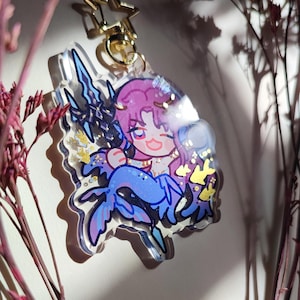 Puede incluir: Un llavero de acr&iacute;lico transparente con un personaje de dibujos animados de pelo rosa y cola de sirena azul, sosteniendo un tridente oscuro. El dije incluye detalles de purpurina dorada en forma de estrella y un cierre de estrella. Texto inferior: ART BY @HSAPHIRYA - SAPHIRYA.STORENVY.COM.