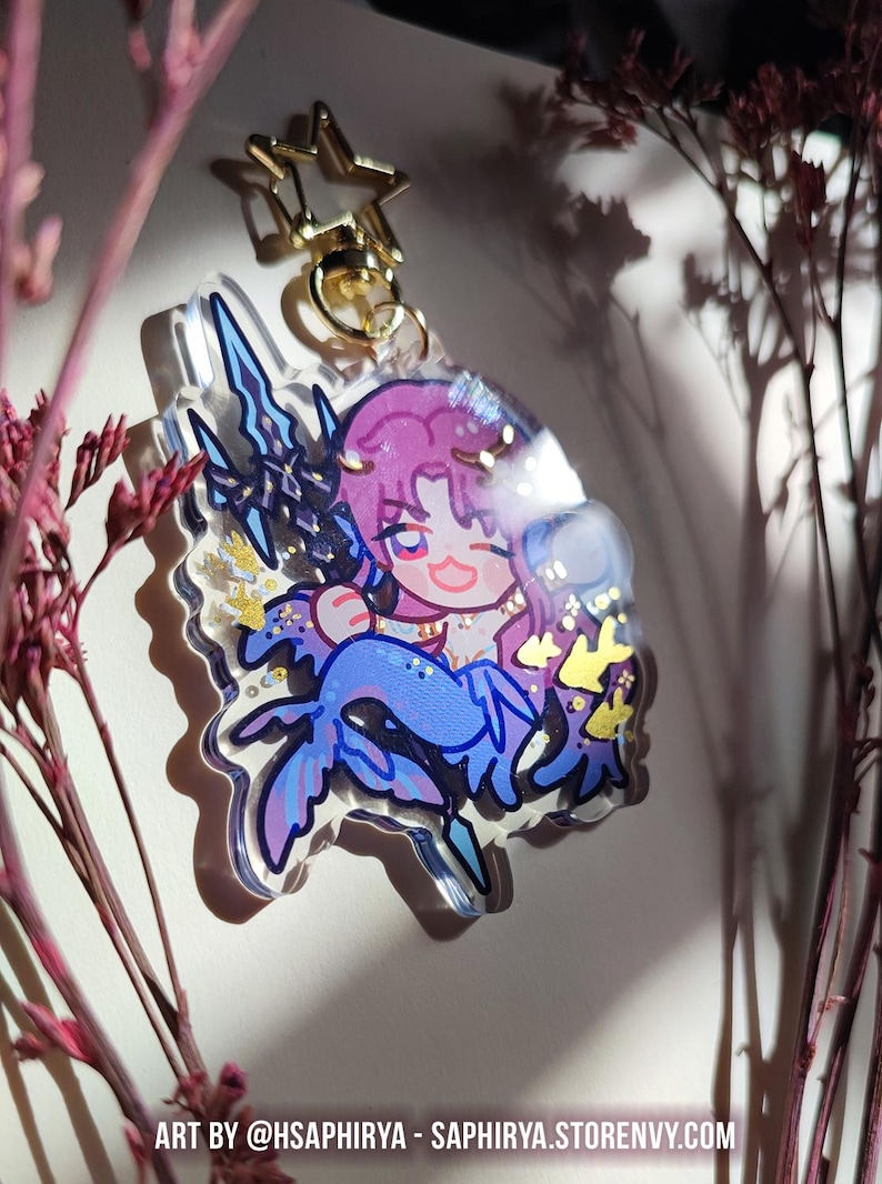 Puede incluir: Un llavero de acr&iacute;lico transparente con un personaje de dibujos animados de pelo rosa y cola de sirena azul, sosteniendo un tridente oscuro. El dije incluye detalles de purpurina dorada en forma de estrella y un cierre de estrella. Texto inferior: ART BY @HSAPHIRYA - SAPHIRYA.STORENVY.COM.