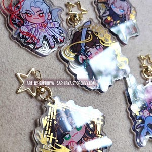 Puede incluir: Una colecci&oacute;n de cinco dijes de acr&iacute;lico transparente con personajes de anime estilo chibi y cierres dorados en forma de estrella. Cada dije muestra ilustraciones &uacute;nicas con colores vibrantes y detalles en l&aacute;mina dorada. Texto: ART BY SAPHIRYA - SAPHIRYA.STORENVY.COM.