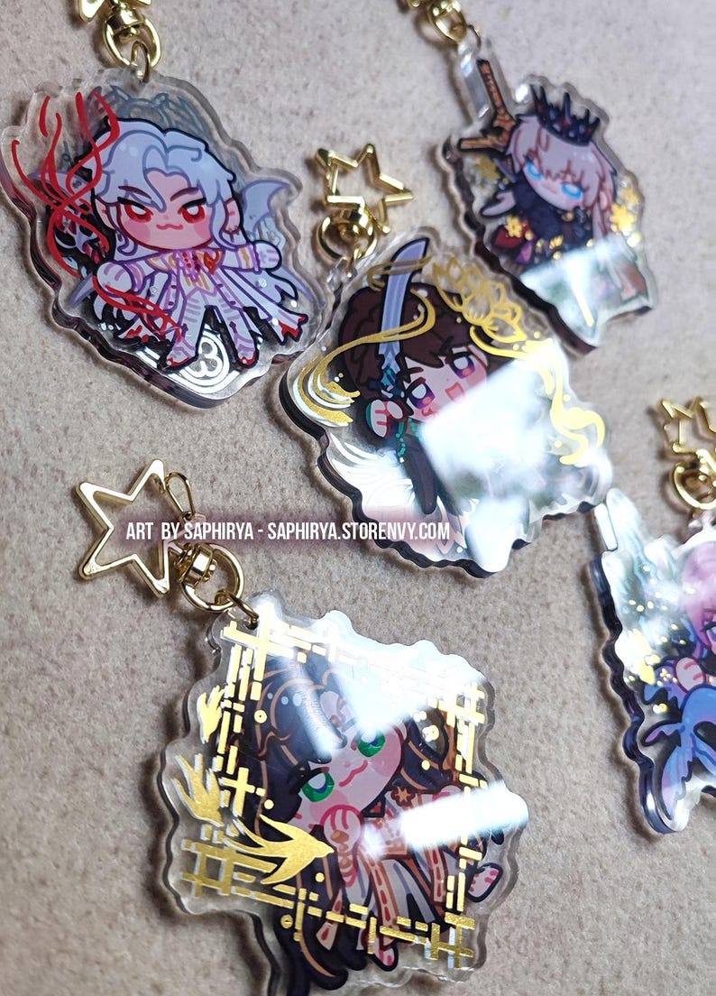 Puede incluir: Una colecci&oacute;n de cinco dijes de acr&iacute;lico transparente con personajes de anime estilo chibi y cierres dorados en forma de estrella. Cada dije muestra ilustraciones &uacute;nicas con colores vibrantes y detalles en l&aacute;mina dorada. Texto: ART BY SAPHIRYA - SAPHIRYA.STORENVY.COM.