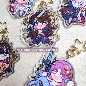Puede incluir: Una colecci&oacute;n de cinco llaveros de acr&iacute;lico transparente con coloridas ilustraciones de personajes estilo anime y cierres dorados en forma de estrella. El texto dice: ART BY SAPHIRYA - SAPHIRYA.STORENVY.COM.