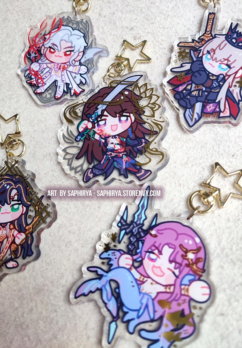 Puede incluir: Una colecci&oacute;n de cinco llaveros de acr&iacute;lico transparente con coloridas ilustraciones de personajes estilo anime y cierres dorados en forma de estrella. El texto dice: ART BY SAPHIRYA - SAPHIRYA.STORENVY.COM.