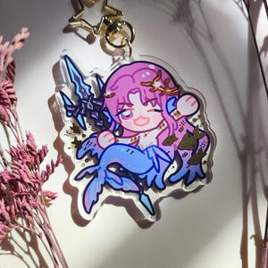 Puede incluir: Un llavero de acr&iacute;lico transparente con un personaje estilo chibi de pelo rosa, cola de sirena azul y una lanza oscura. El personaje lleva una corona dorada y est&aacute; rodeado de detalles en pan de oro. Incluye un cierre en forma de estrella dorada.
