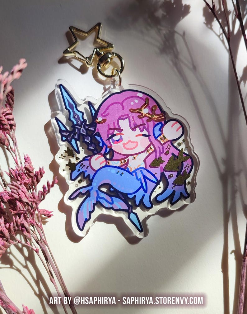 Puede incluir: Un llavero de acr&iacute;lico transparente con un personaje estilo chibi de pelo rosa, cola de sirena azul y una lanza oscura. El personaje lleva una corona dorada y est&aacute; rodeado de detalles en pan de oro. Incluye un cierre en forma de estrella dorada.